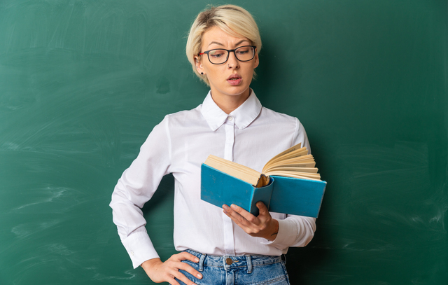 impressed young blonde female teacher wearing glasses in classroom standing in front of chalkboard holding and reading book keeping hand on waist